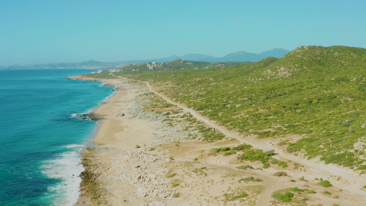 Laguna Beach Los Cabos aerial frame with surf breaking along the left edge, pale sand and rocks in the lower middle, intersecting dirt paths near the beach, green hills to the right, and no visible people.