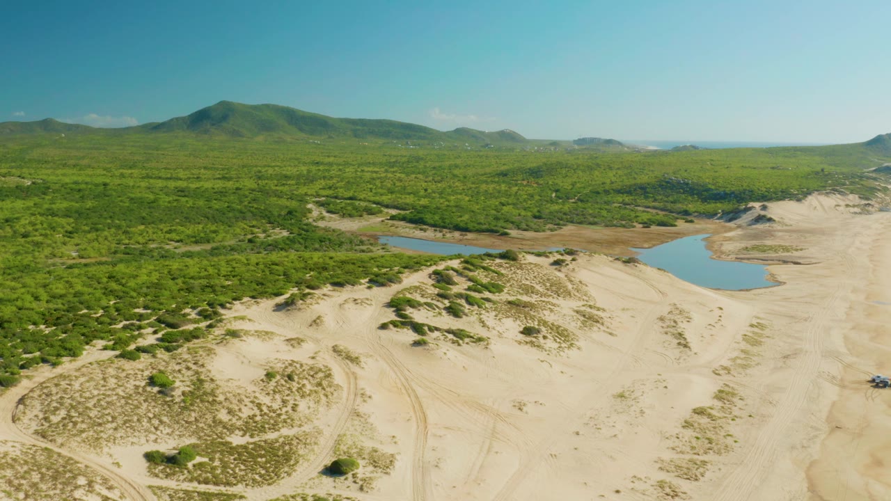 Aerial frame from Laguna Beach Los Cabos showing pale dunes in the foreground, two small blue water bodies near the green vegetation, distant hills, and no visible people.