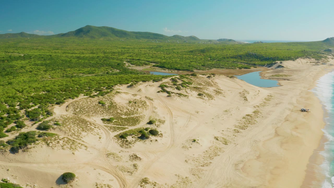 Aerial frame at 0:01 over Laguna Beach Los Cabos with the dune ridge centered, lagoon pools behind it, the surf line on the right edge, forest and hills beyond, and no visible people.