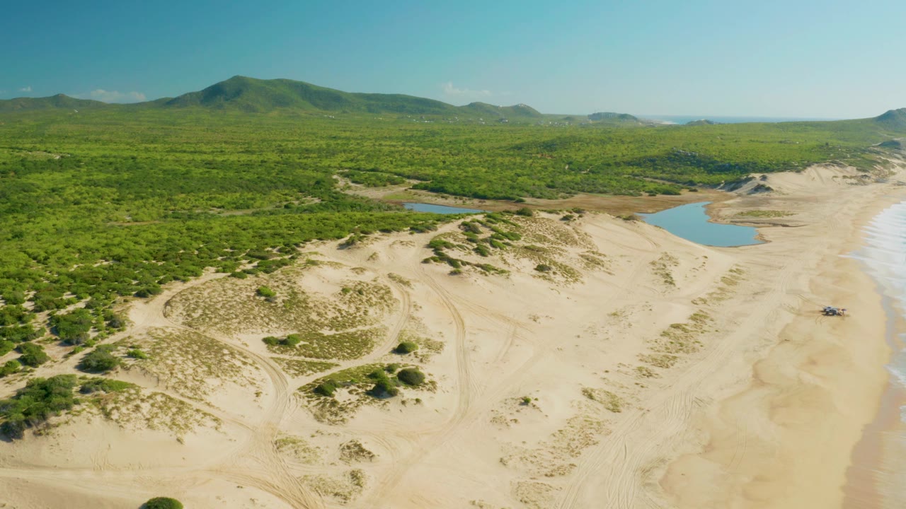 Aerial frame at 0:02 shows Laguna Beach Los Cabos from above with more surf visible along the right side, broad pale sand, lagoon water behind the dunes, green forest, distant hills, and no visible people.