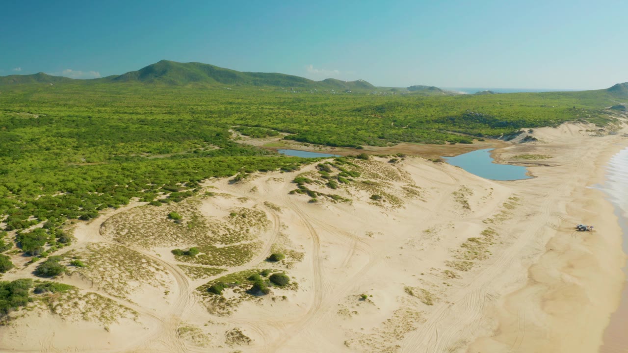 Mid-clip aerial frame over Laguna Beach Los Cabos with the vehicle visible near the right beach area, pale dunes in front, lagoon pools behind, green forest across the horizon, and no visible people.