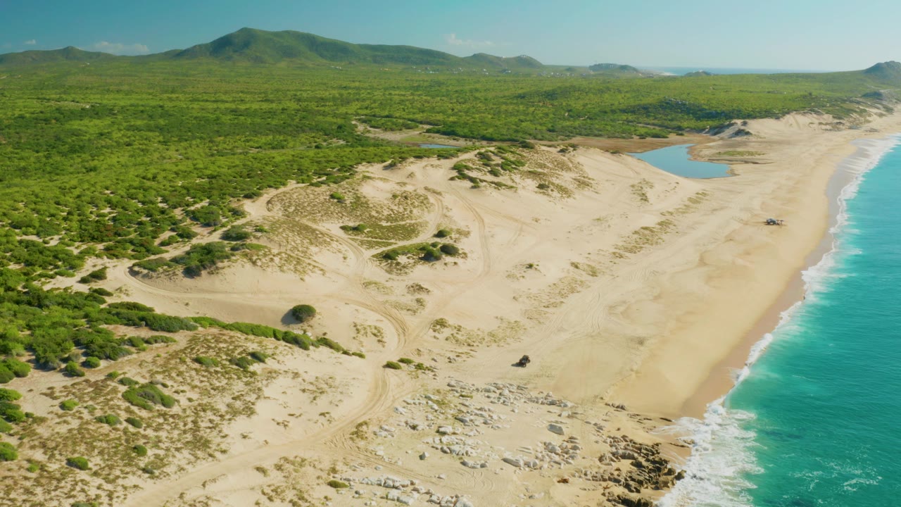 Aerial view of sand dunes, green vegetation, turquoise surf, and distant hills.
