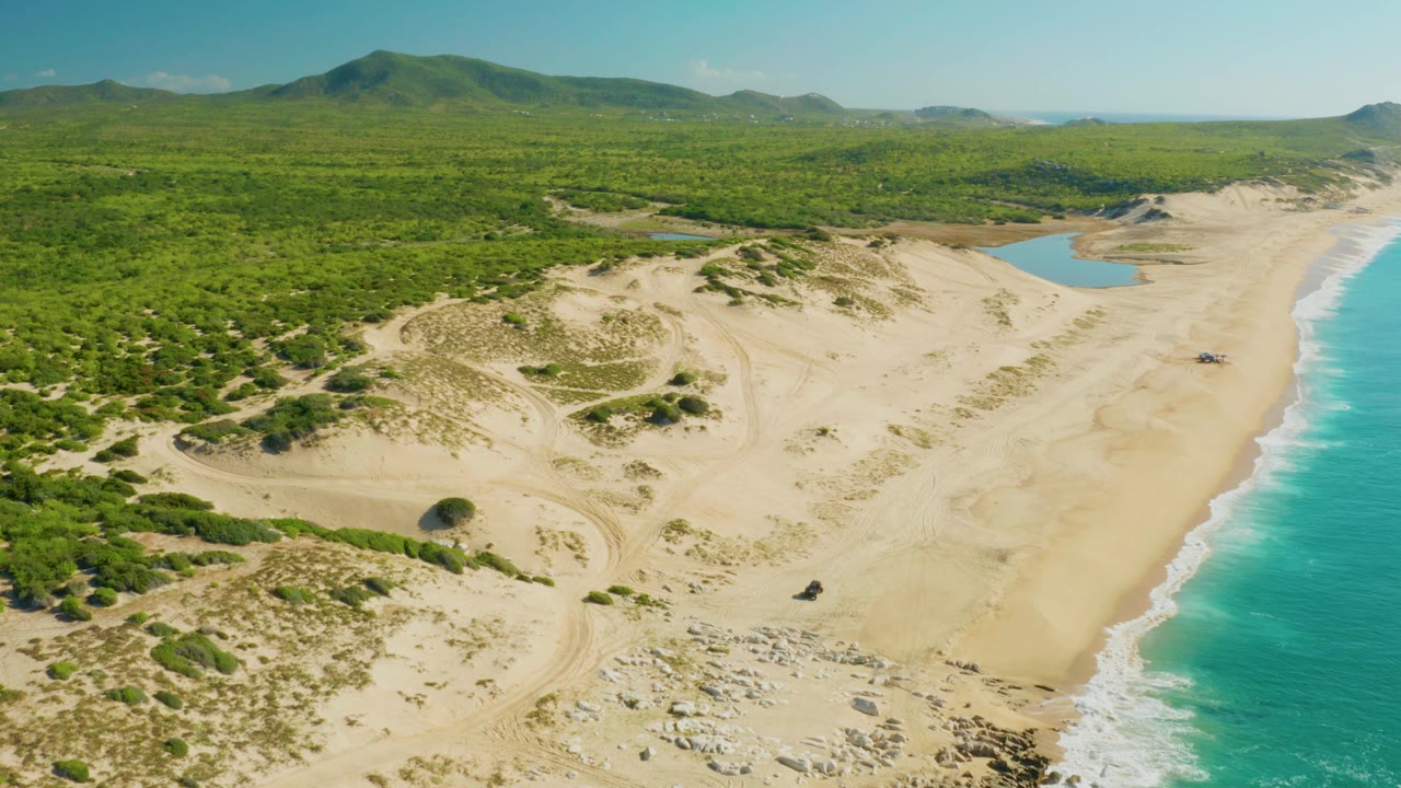 Aerial frame from Laguna Beach Los Cabos with the camera looking across sand tracks and dunes toward a lagoon, forested green terrain, turquoise shoreline, and no visible people.