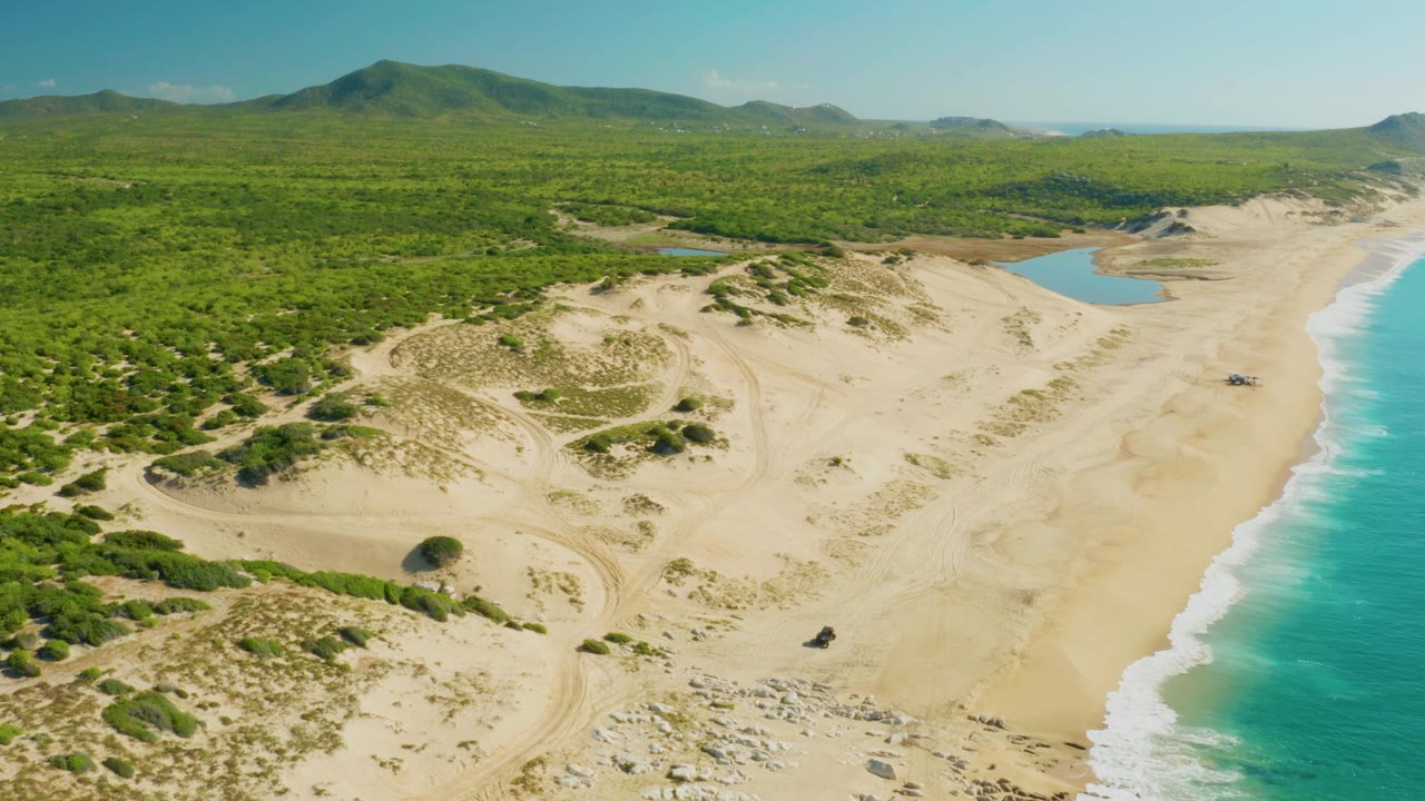 Wide aerial view over Laguna Beach Los Cabos with scrub-covered dunes filling the left side, a sandy beach and lagoon in the center, ocean surf at right, and no visible people.