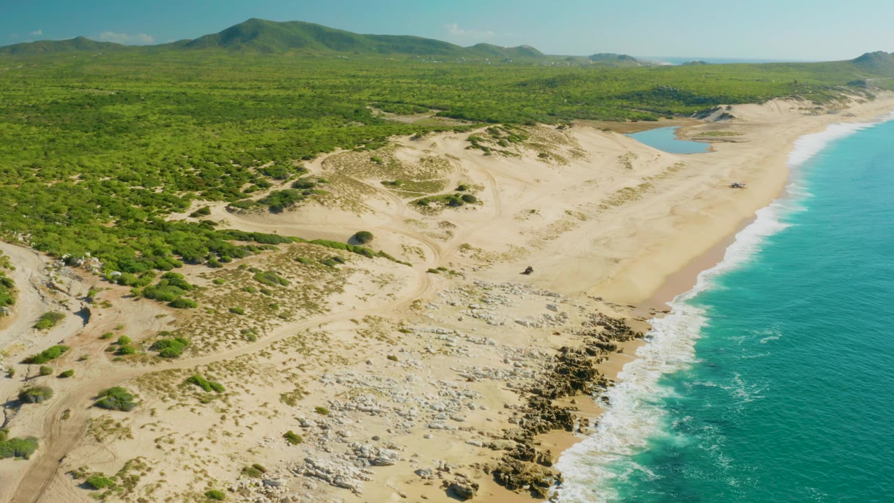 Aerial view of sand dunes, green scrub, rocky shore, and turquoise ocean.