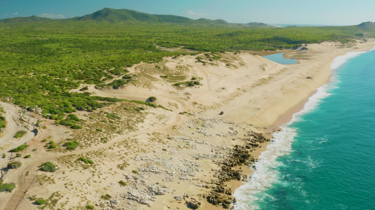 Wide aerial view at Laguna Beach Los Cabos with the beach and surf line shifted slightly right, dune tracks and green scrub visible inland, distant hills, and no visible people.