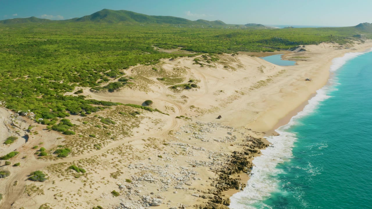 Aerial frame from Laguna Beach Los Cabos showing a longer stretch of turquoise water at right, pale sand dunes in the center, green vegetation behind them, distant hills, and no visible people.