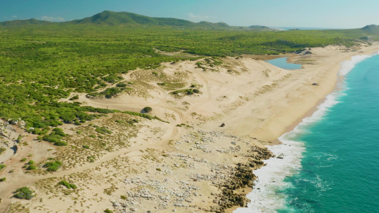 Drone-style aerial view over Laguna Beach Los Cabos with scrub-covered dunes in the foreground, a rocky shoreline below, ocean surf along the right edge, distant hills, and no visible people.