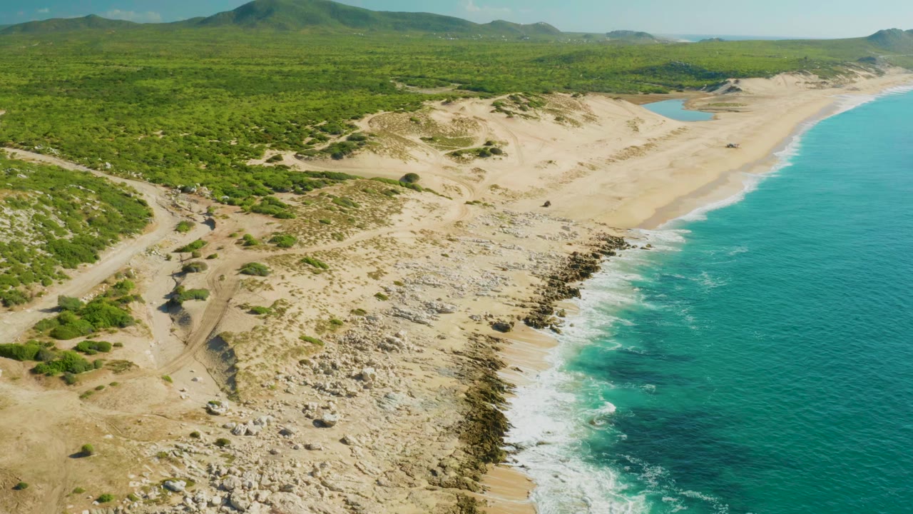 Aerial frame over Laguna Beach Los Cabos where the rocky center shoreline leads toward a wide sandy beach, with turquoise surf to the right and no visible people.