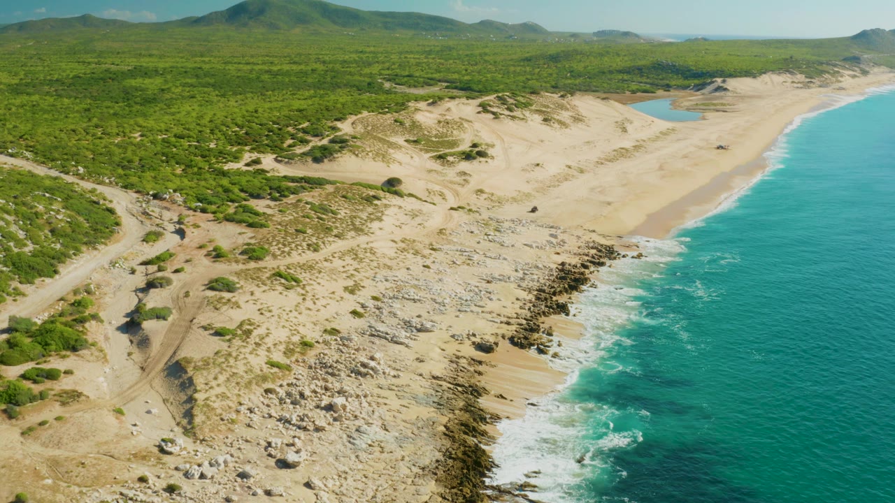 Aerial coastal view in Laguna Beach Los Cabos with a rocky sand strip in the foreground, a broad beach beyond it, green terrain inland, turquoise surf at right, and no visible people.