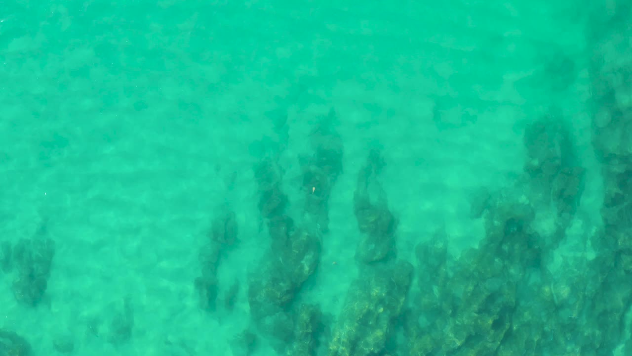 Mid-clip frame from Laguna Beach Los Cabos showing clear turquoise water, sandy bottom, and darker vegetation gathered toward the lower part of the view, with no visible people.