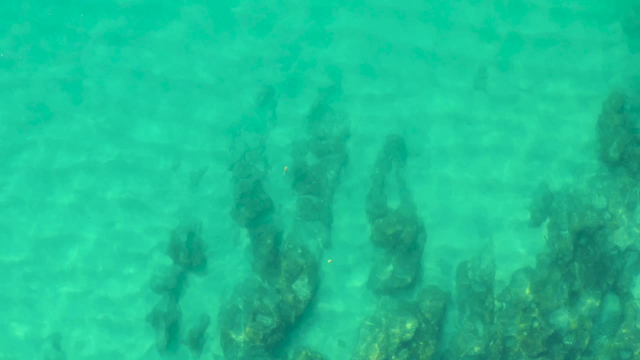 Shallow turquoise water passes over darker seabed patches and soft sandy-toned areas at Laguna Beach Los Cabos, with no visible people.