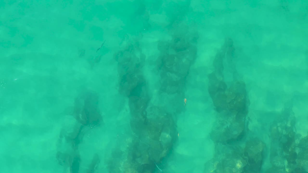 Overhead view at Laguna Beach Los Cabos where turquoise water reveals reef-like submerged rocks and faint sandy areas, with no visible people.