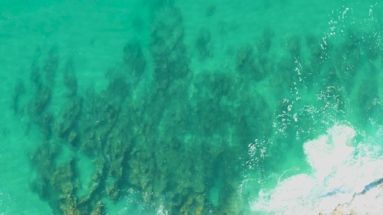 Clear turquoise sea at Laguna Beach Los Cabos reveals long dark rock shapes under the surface, while white surf foam gathers along the right side with no visible people.