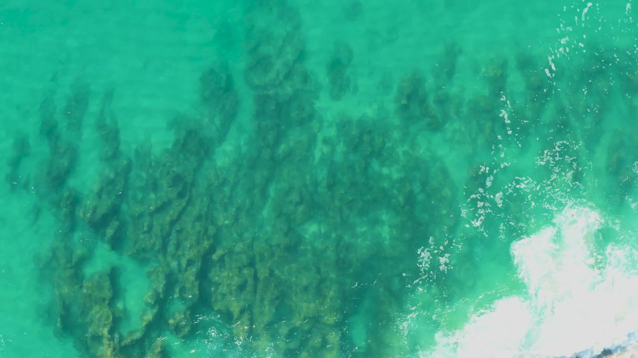 Aerial view over green-blue shallow water at Laguna Beach Los Cabos, with clustered underwater rocks centered in frame and a patch of foam on the right, no visible people.