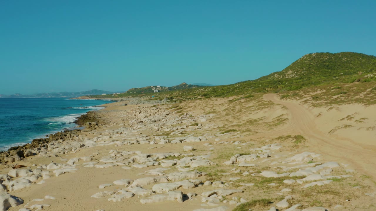 Rocky sandy beach beside blue ocean and a green hill under clear sky.