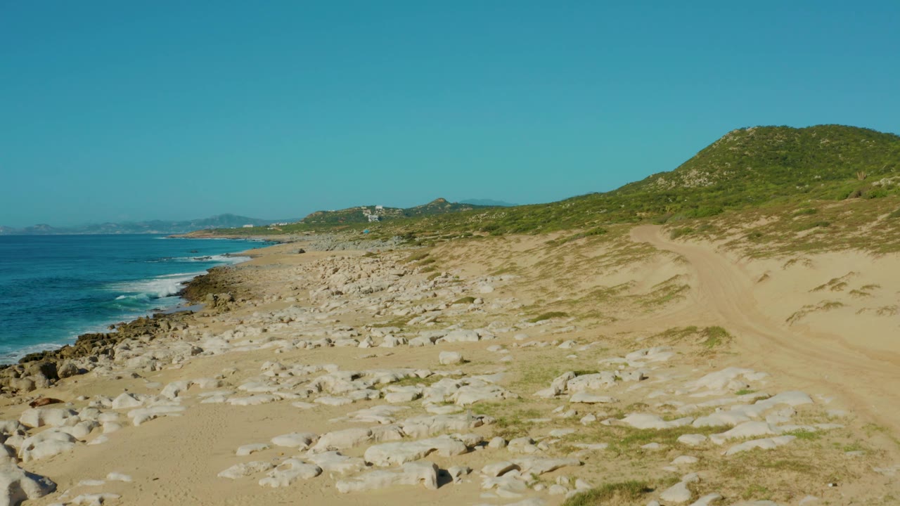 Laguna Beach Los Cabos shoreline frame with surf breaking along the left edge, scattered white rocks on sand, a green hill to the right, and no visible people.
