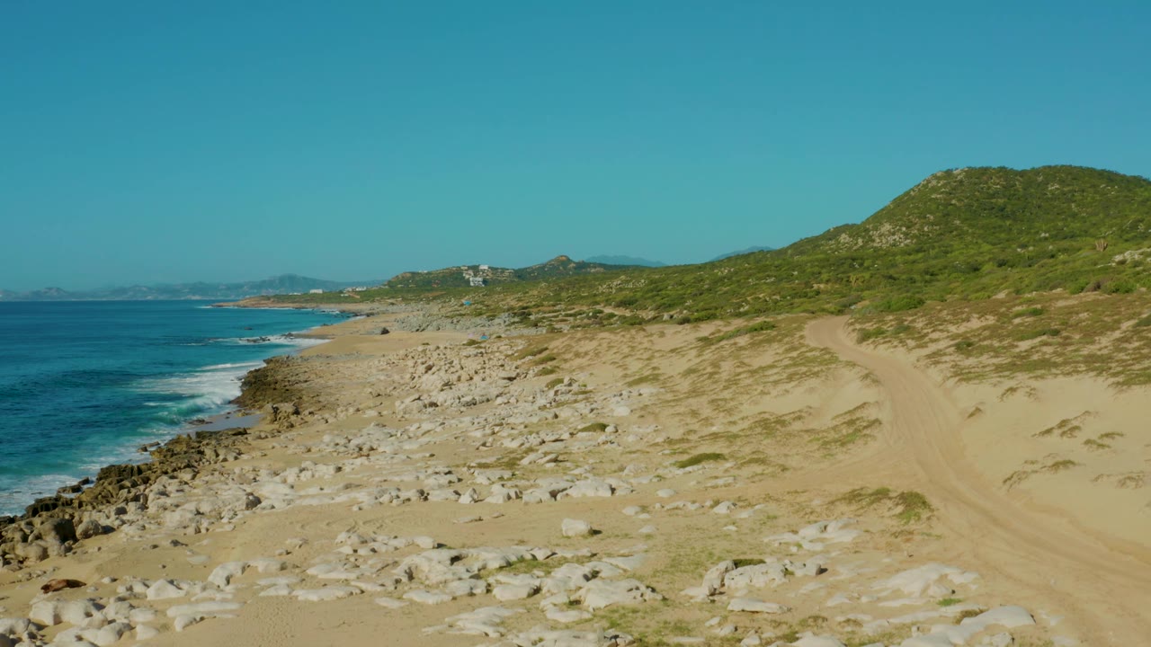 Laguna Beach Los Cabos view with turquoise water and white surf at left, a broad field of rocks and sand in the middle, green hillside at right, and no visible people.