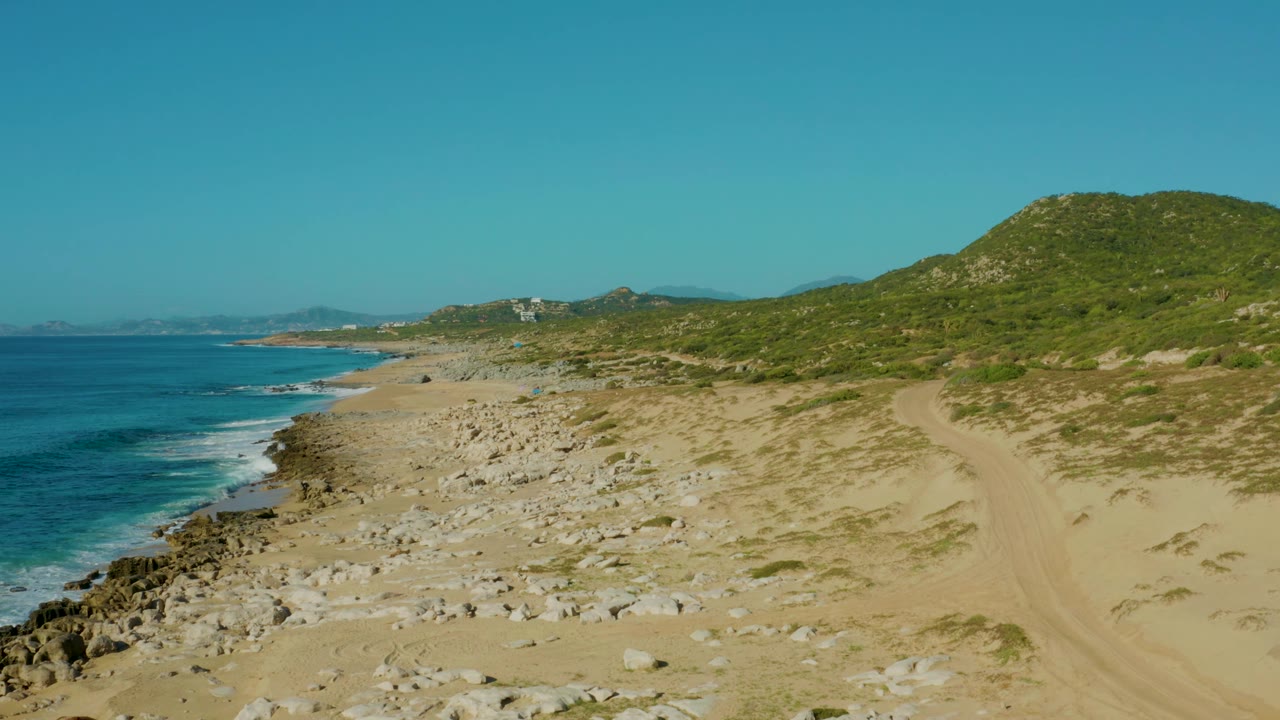 Mid-clip frame from Laguna Beach Los Cabos showing the ocean farther left, rocky sand across the beach, faint sand tracks near the hillside, green vegetation, and no visible people.