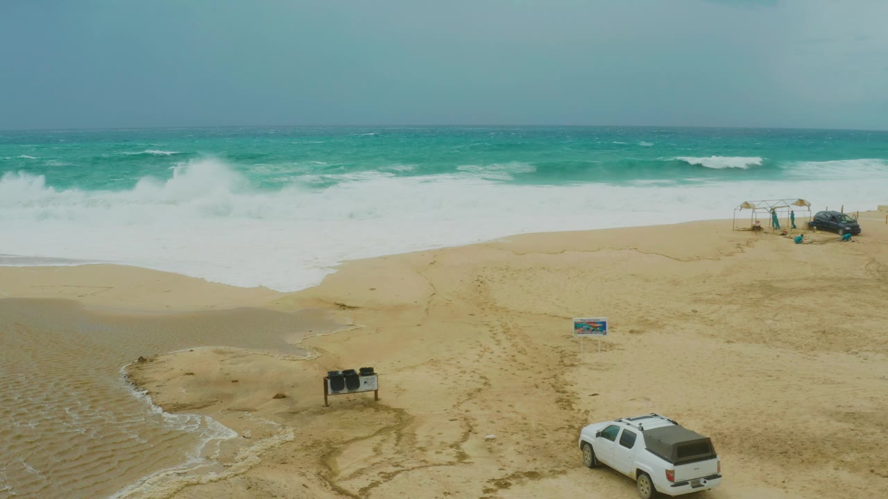 Aerial frame over Dreams Resort Los Cabos beach showing white surf rolling toward sand, a shallow water channel, a white vehicle near the lower right, small beach structures, and no visible people.