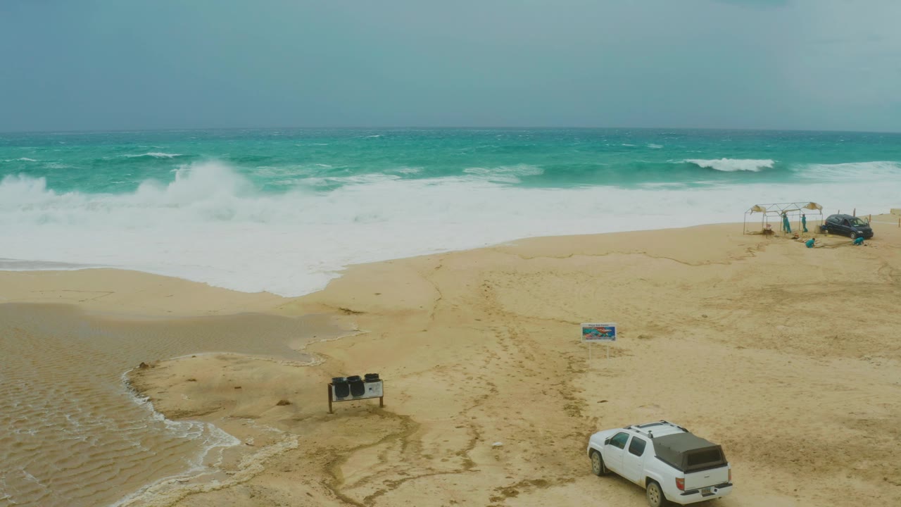 Aerial view at Dreams Resort Los Cabos with the white vehicle farther along the lower edge, foamy waves spread across the upper beach, and no visible people near the small structures.