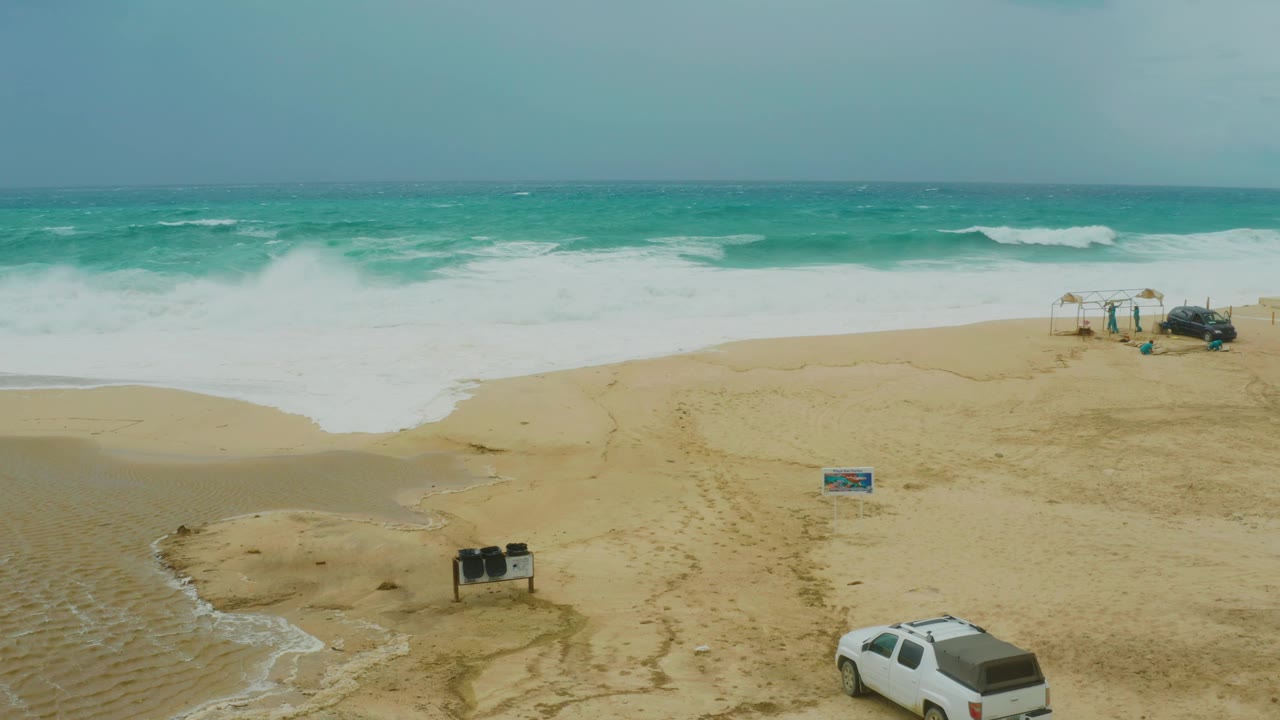 Dreams Resort Los Cabos beach from above, with a broad band of white surf, sand tracks across the beach, the white vehicle near the lower right edge, and no visible people.