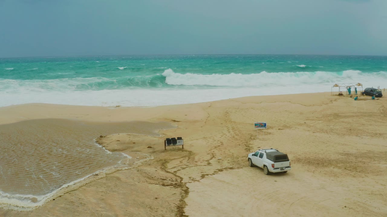 One second in, the Dreams Resort Los Cabos shoreline remains in an elevated composition, with the white SUV slightly farther along the lower right sand and broad white surf breaking across the background.