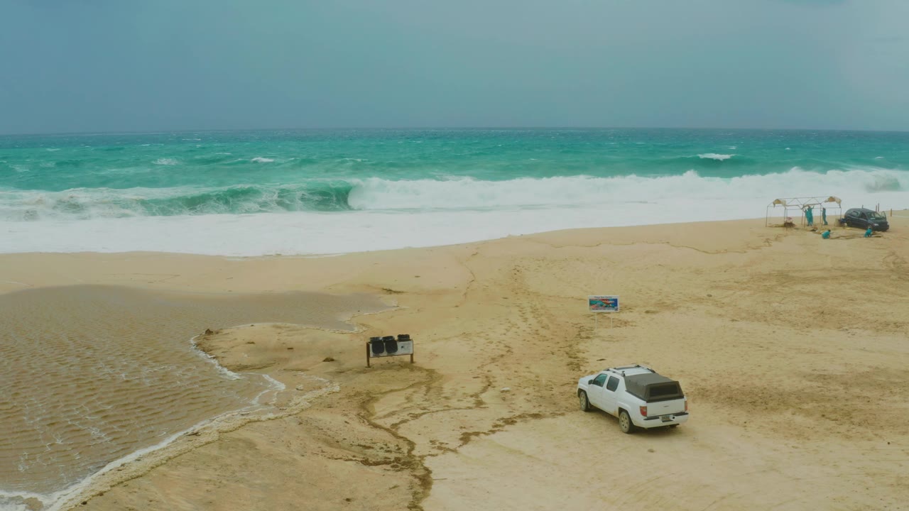 At the midpoint, overcast light flattens the tan beach and blue-green ocean while the white SUV sits lower right and foamy surf spreads wider across the upper shoreline.