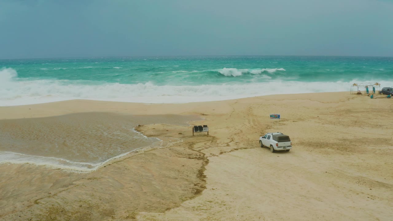 White SUV drives across sand below breaking waves