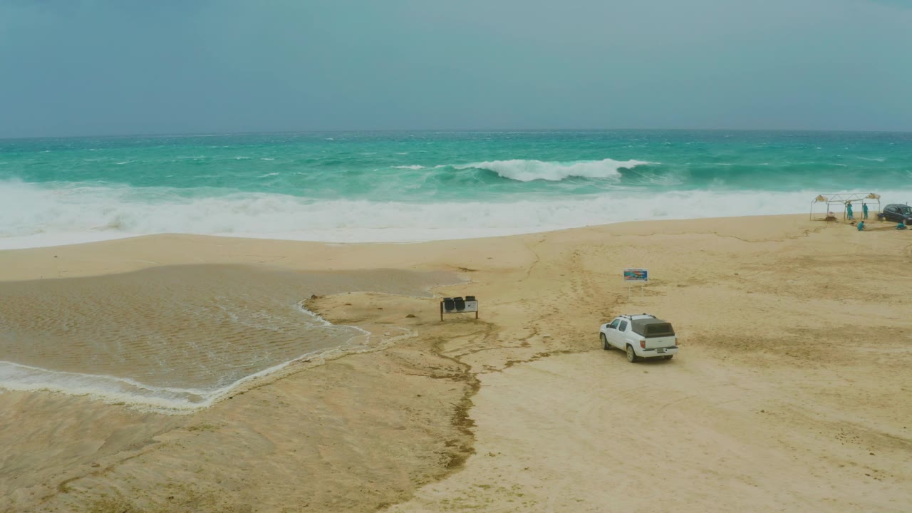 The white SUV remains in the lower right of the Dreams Resort Los Cabos beach frame, with darker tire marks leading toward beach fixtures and waves rolling in behind them.