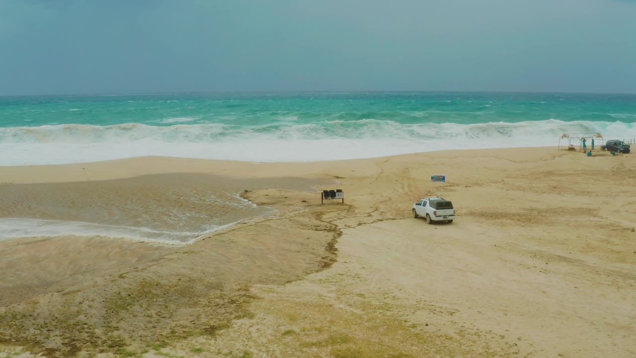 Aerial-style view at Dreams Resort Los Cabos showing the parked white SUV below a broad band of turquoise water and white breakers, with no visible people.