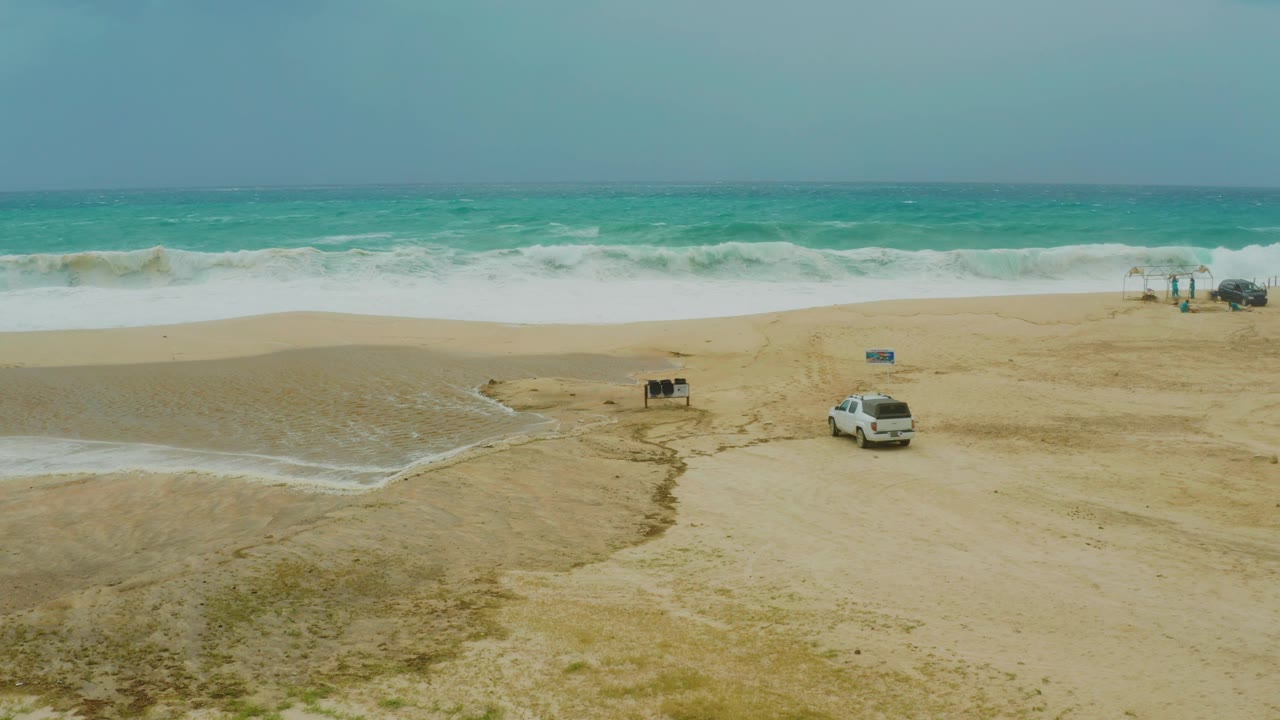 Mid-clip frame at Dreams Resort Los Cabos with crashing waves, a parked white SUV on open sand, small beach structures, and no visible people.