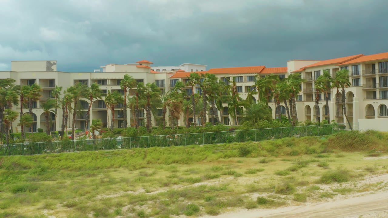 Opening frame of Dreams Resort Los Cabos in Baja California Sur, Mexico, showing the hotel behind palms, a green fence, dune grass, and no visible people.