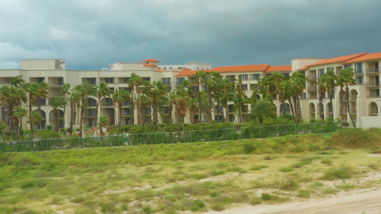 The resort remains in view with orange roof sections, clustered palms, a low green fence, pale dune sand, and no visible people in the frame.