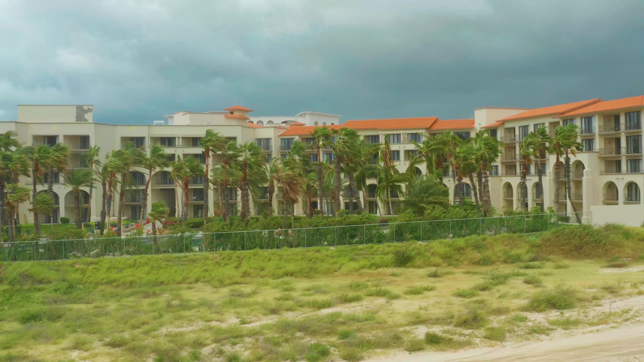 Resort buildings, palms, fence, dunes, and cloudy sky.