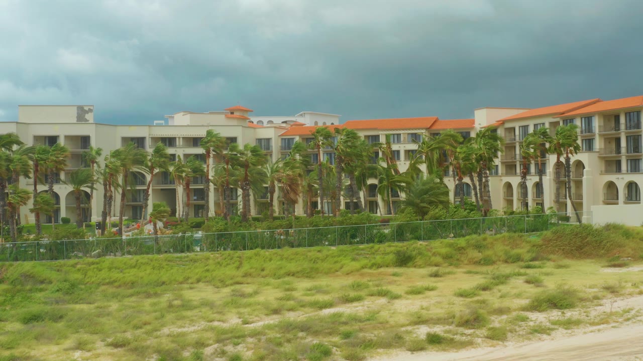 A palm-lined section of the Dreams Los Cabos resort sits beyond patchy sand and grass, with small human figures difficult to distinguish near the fence area.
