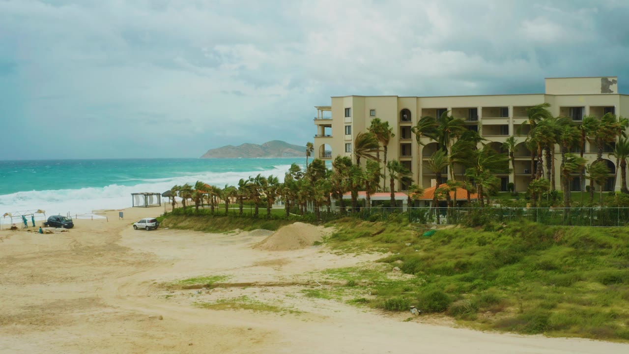 Opening frame at Dreams Resort Los Cabos shows sandy beach in front, turquoise surf at left, a multi-story coastal building at right, palm trees, vehicles, and small people near the shoreline.