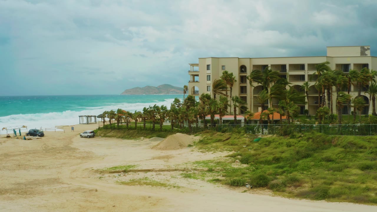 Sandy resort beach with ocean surf, vehicles, palms, and coastal building