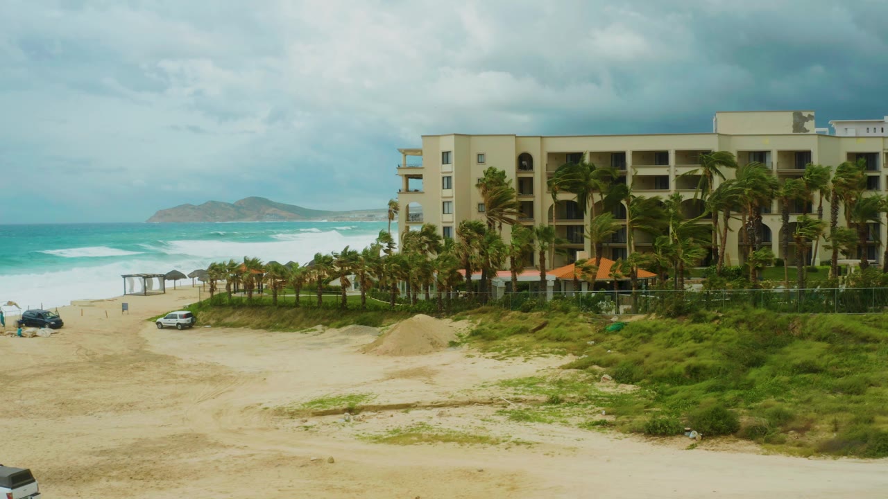 Wide beachfront resort with palm trees, sandy beach, ocean waves, and two cars.