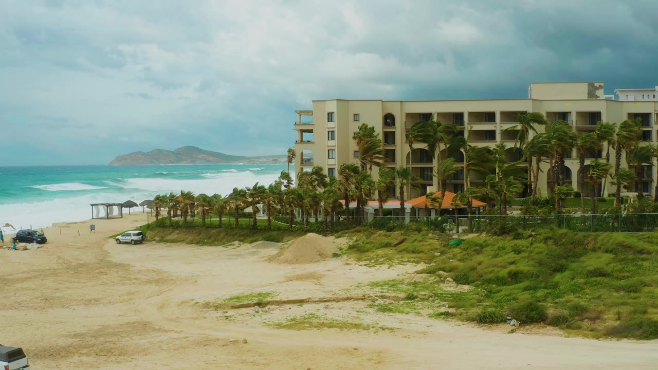 Dreams Resort Los Cabos remains on the right side of frame as waves break on the left, with two cars positioned on the sandy beach and no visible people.