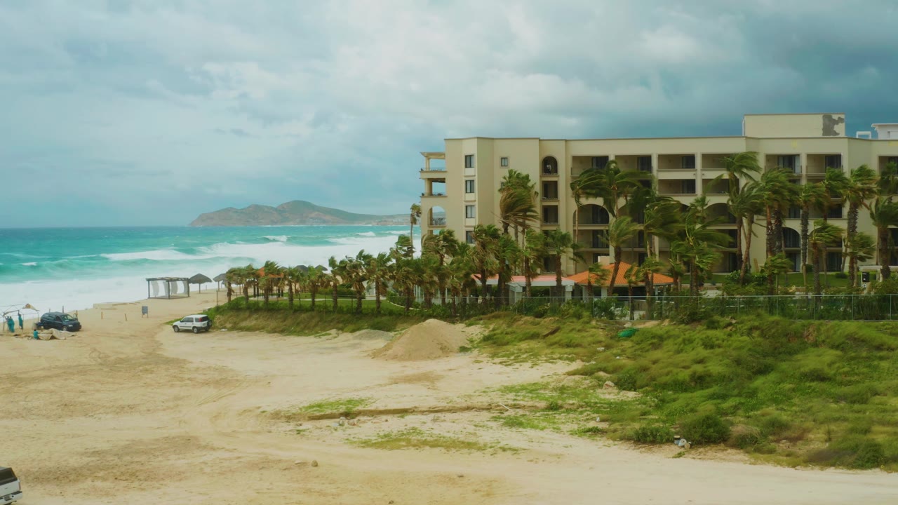 Mid-clip frame of Dreams Resort Los Cabos with the building filling the right side, a row of palms across the beach, two cars on the sand near the surf, and no visible people.