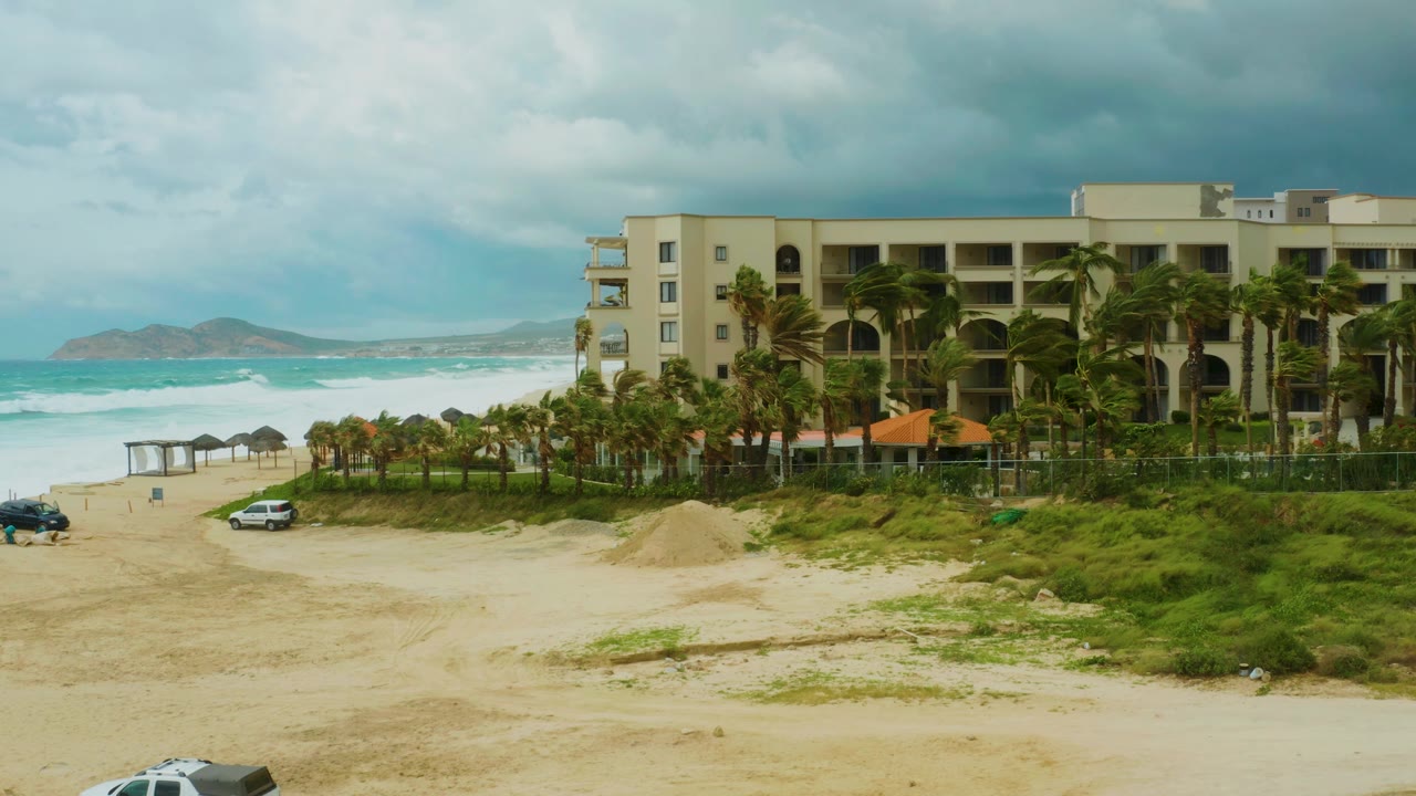 At Dreams Resort Los Cabos, the white car is farther along the sandy beach path below palm trees, a beige resort facade, choppy turquoise waves, distant land, and no visible people.