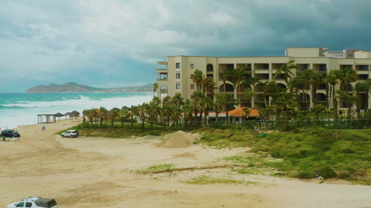 At Dreams Resort Los Cabos, the white car continues across the sand beneath a cloudy sky, with palms in front of the resort building and ocean waves rolling in behind, no visible people.