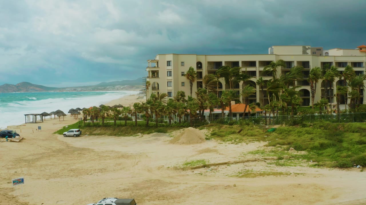 At 0:01, the white car sits farther along the sandy road below the palm-lined Dreams Resort Los Cabos exterior, with waves and distant hills visible to the left and no visible people.