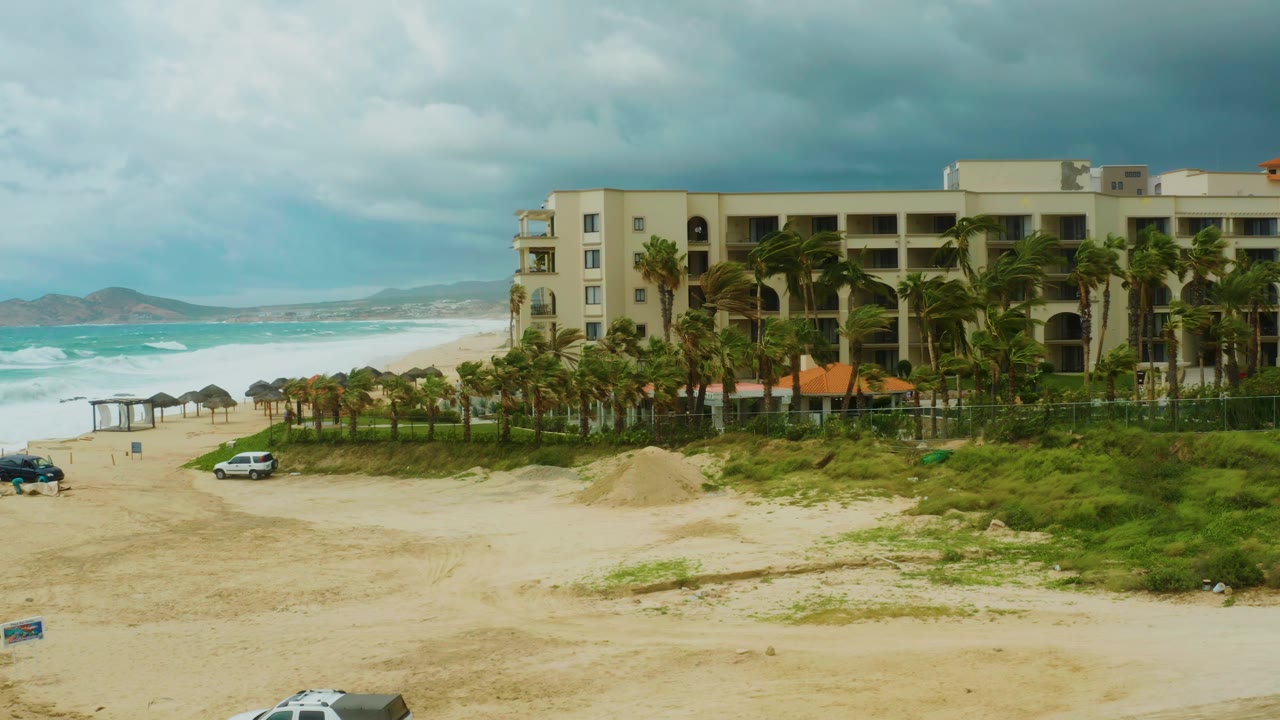 Midway through the clip, the white car is visible on the sandy road below the hotel and palms at Dreams Resort Los Cabos, with the ocean and low hills on the left and no visible people.