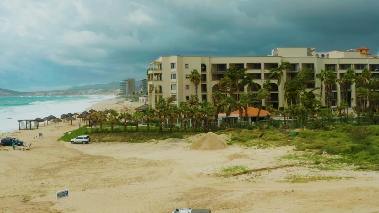 Dreams Resort Los Cabos remains centered beside the beach as waves roll in at left, with a white vehicle on the sand, beach umbrellas, a sand pile, and no visible people.