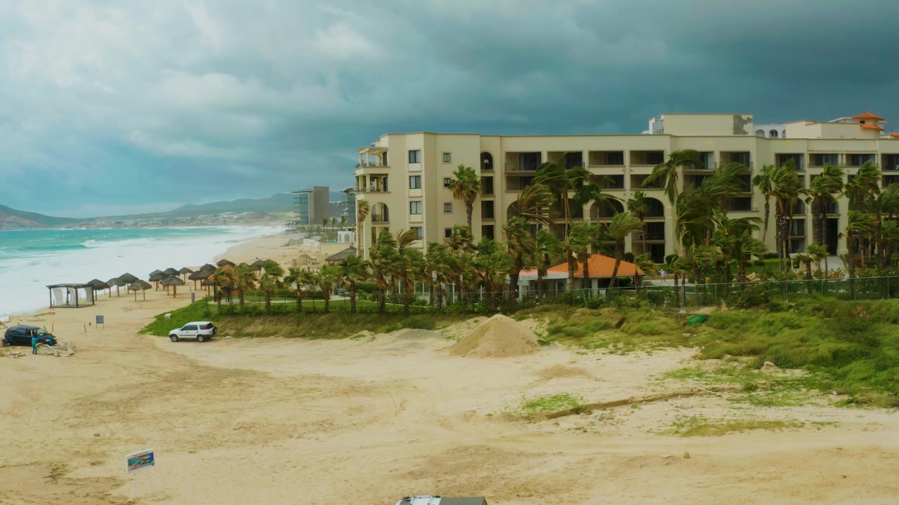 A wide Dreams Resort Los Cabos frame shows the hotel on the right, turquoise waves on the left, beach umbrellas near the water, vehicles on the sand, and no visible people.