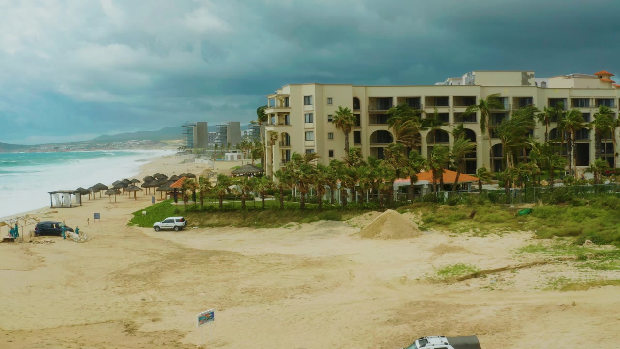 Elevated view of Dreams Resort Los Cabos with a sandy beach, turquoise surf, palm trees, vehicles on the sand, and small visible people near the shoreline at the start of the clip.