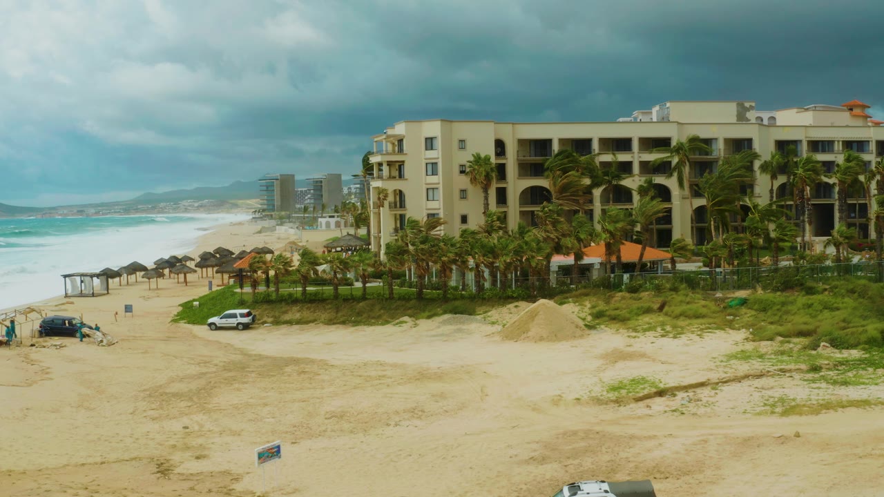 A wide elevated frame shows the Dreams Resort Los Cabos beachfront building, wind-leaning palms, choppy ocean water, sandy foreground, and small people and vehicles near the beach.