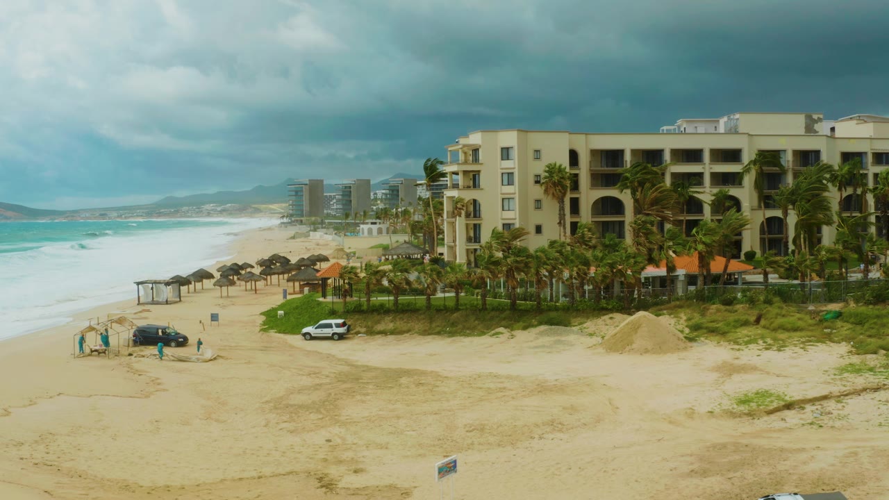 Wide elevated view of Dreams Resort Los Cabos with a sandy beach, small shade structure, parked cars, palm trees, surf, distant buildings, and tiny visible people under dark clouds.